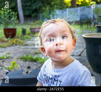 A grubby small caucasian boy (age 11 months) with blue eyes wearing a soiled white t-shirt top sits in the garden with a quizzical expression Stock Photo