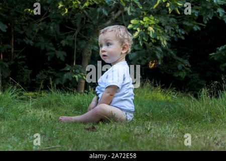 A small caucasian boy (age 11 months) with blue eyes wearing a white t-shirt top sits on grass in the garden with a quizzical expression Stock Photo
