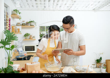 Young asian man and woman together cooking cake and bread with egg, looking menu from tablet in the flour happy relaxing in at home Stock Photo