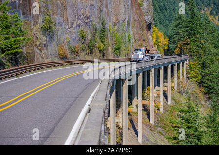 Day cab white big rig powerful American bonnet semi truck transporting fixed metal pipes on flat bed semi trailer running on the bridge on the side of Stock Photo