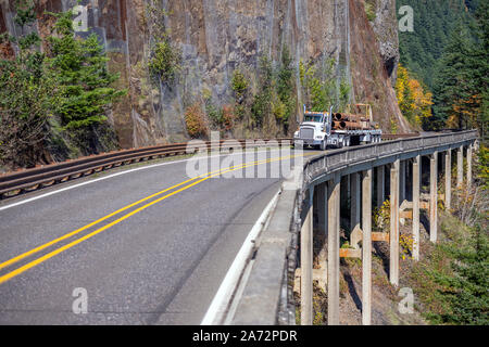 Day cab white big rig powerful American bonnet semi truck transporting fixed metal pipes on flat bed semi trailer running on the bridge on the side of Stock Photo