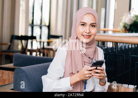 Optimistic Asian woman in hijab smiling and looking at camera while sitting in cafe and browsing social media on mobile phone Stock Photo