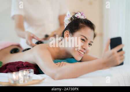 Beautiful young Asian woman smile and holding smartphone receiving rock therapy massage taking selfie in spa salon. Hand putting rock on female back, Stock Photo