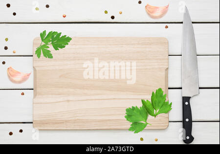 Rustic culinary template - top view of an empty cutting board with spices ( garlic, pepper, parsley) and knife on a white wooden table. Stock Photo