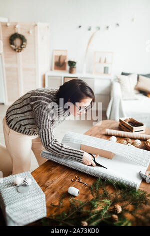 Young woman preparing Christmas presents for holiday she cutting wrapping paper and wrapping them at the table Stock Photo