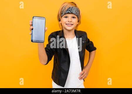 naughty handsome boy in a black jacket and bandana holds the phone with the screen forward, yellow isolated background Stock Photo