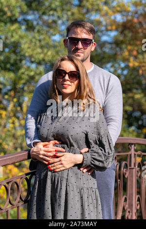 Loving young couple posing on a wrought iron bridge outdoors in an autumn park standing in a close embrace smiling at the camera Stock Photo