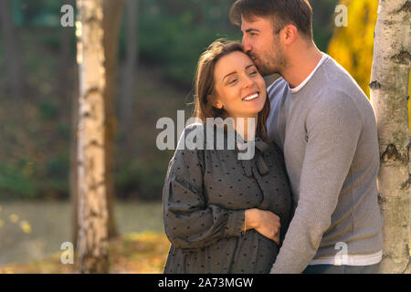 Happy young pregnant woman cuddling with her husband as they lean against a tree in a park in evening light Stock Photo