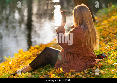 Young woman posing for a selfie in an autumn park as she sits relaxing on the grass overlooking water with reflections of sunlight Stock Photo
