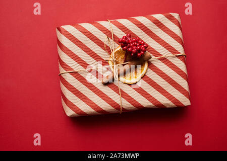 Close-up of Christmas gift box in striped wrapping paper decorated with dried berries and cinnamon isolated on red background Stock Photo
