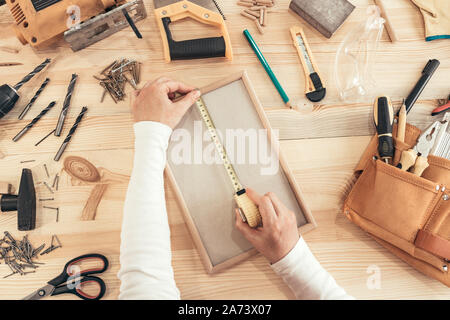 Female carpenter tape measuring picture frame in small business woodwork workshop, top view of hands Stock Photo