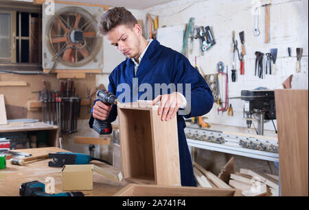 Young positive woodworker using drilling machine to make drawer at workshop Stock Photo