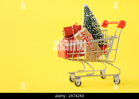 Shopping cart full of various gift boxes and a Christmas tree on yellow background. Christmas sale concept. Stock Photo