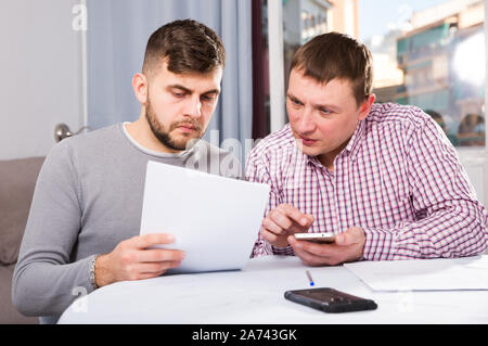 Two serious male friends reading attentively and discussing documents, using phone at home table Stock Photo