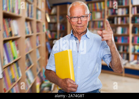 old-aged man in glasses standing in bookstore with book in his hands, pointing with his index finger up, strongly recommending reading Stock Photo