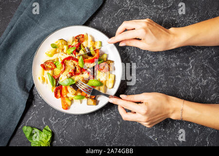Female hands hold knife and fork. Pasta with grilled vegetables - zucchini, eggplant, pepper ant tomato on black stone background. Top view Stock Photo
