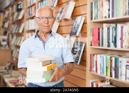 Elderly male holding stack of books near bookshelves in bookshop interior Stock Photo