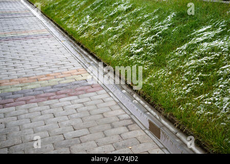 Wet tiled sidewalk at the beginning of winter. Lawn with green grass covered in snow. A gutter separates the lawn and the pedestrian walkway. Stock Photo