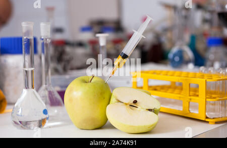 Syringe with chemical additive in fresh apple on background with laboratory equipments. Concept of research of food genetic modification Stock Photo