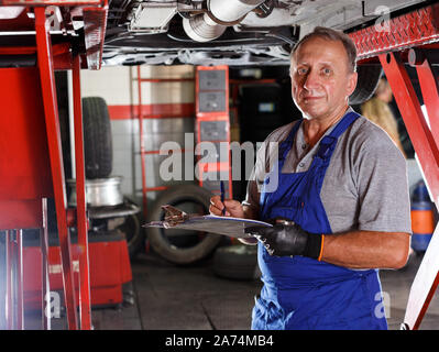 Skilled mature male mechanic taking notes in notebook about car repair at auto service Stock Photo