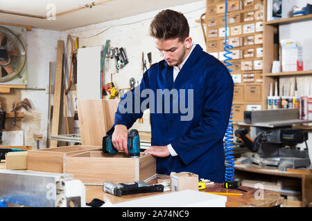Young positive man using drilling machine to make drawer at workshop Stock Photo