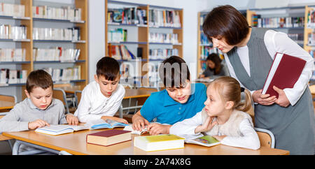 Group of happy positive school kids studying in school library with friendly female teacher Stock Photo
