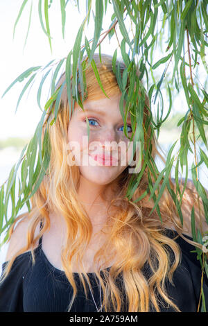Portrait of a gorgeous smiling teenage girl with red hair outdoors hiding behind leaves Stock Photo