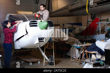 Two young happy positive smiling mechanics, man and woman, repairing light sport plane in workshop Stock Photo