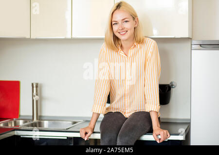 Young lovely blonde woman posing sitting on kitchen cabinet near table with fresh organic vegetables and fruits, smiling looking at camera. Healthy fo