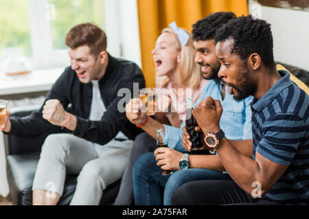 Side view shot of young african bearded man watching sport match with friends, rooting for favorite team, clenching fists, sitting with drinks on couc Stock Photo