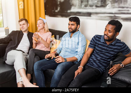 Young handsome bearded african american guy watching interesting exciting movie on tv at home with his multiethnic friends, drinking soft beverages. S Stock Photo