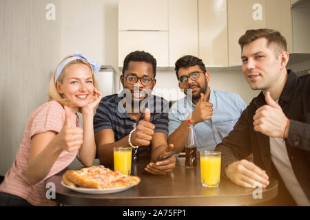 Group of diverse friends sitting at table in kitchen showing thumb up gesture, eating tasty pizza, drinking juice and beer, smiling looking at camera. Stock Photo