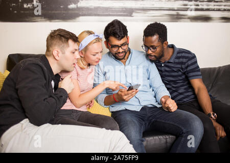 Group of four multiracial friends having fun at home together. Three men and woman using smartphone, watching funny video. Friendship, leisure and tec Stock Photo