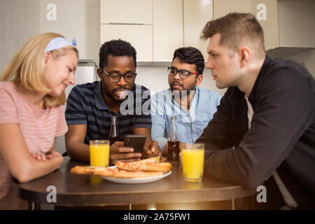 Group of diverse friends sitting at table, eating pizza, drinking juice, using mobile phone, friendly talking. Multicultural students having lunch in Stock Photo