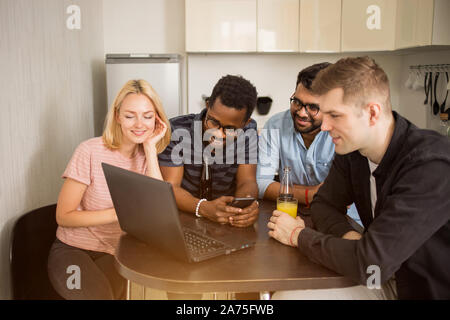 Group of diverse students sitting at table in kitchen, drinking juice, using mobile phone and laptop watching movie together. Friendship, leisure, tec Stock Photo