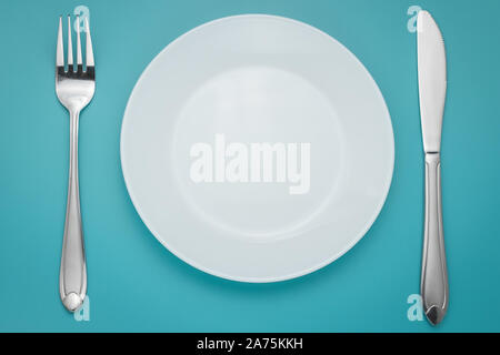 White empty plate with fork and knife on blue background. Turquoise backdrop. Laying, table appointments. Stock Photo