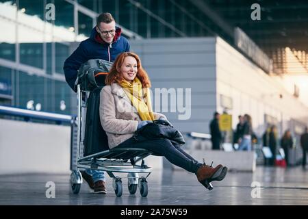Cheerful couple travel together. Young man pushing woman on luggage trolley at airport. Stock Photo