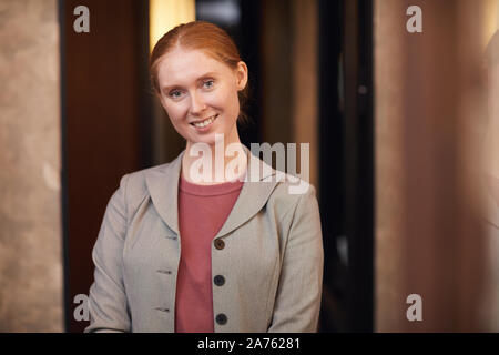 Portrait of red haired young businesswoman smiling at camera while standing at corridor office Stock Photo