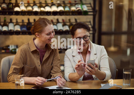 Two smiling businesswomen sitting at the table using mobile phone and discussing work together at the restaurant Stock Photo