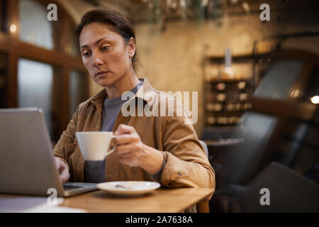 Mature woman drinking coffee and working online on laptop computer at the table in cafe Stock Photo