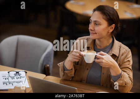 Mature woman drinking coffee while sitting at the table and working on laptop and with documents in coffee shop Stock Photo