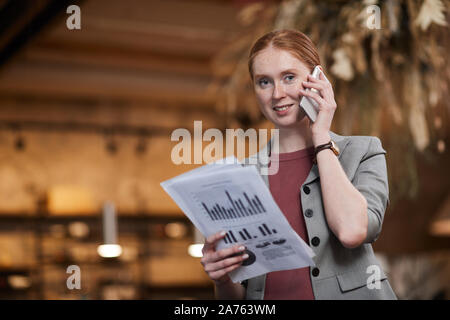 Portrait of red haired businesswoman standing and smiling at camera while has a conversation on mobile phone and examining financial documents Stock Photo
