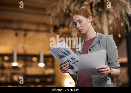 Young red haired businesswoman standing and examining financial documents with charts and graphs Stock Photo