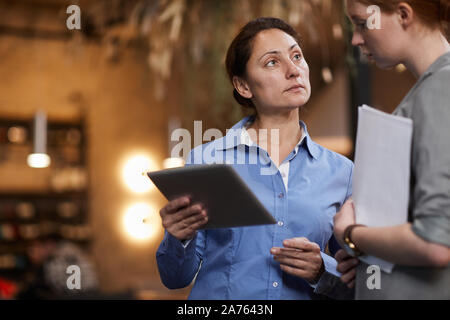 Mature businesswoman holding digital tablet and discussing with her partner online work while they standing in the restaurant Stock Photo