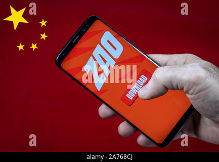 Male hand holding smartfone with ZAO app logo and download button against China flag. Stock Photo
