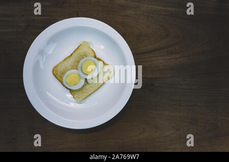 Half cutted hard boiled egg on toasted sandwich bread, ready to eat, in white plate, wooden table background. Vintage toned Stock Photo