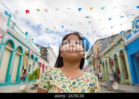 Marechal Deodoro, Alagoas, Brazil - June 21, 2016: Cute little Brazilian girl smiling and looking up to the colorful decoration of pennants Stock Photo