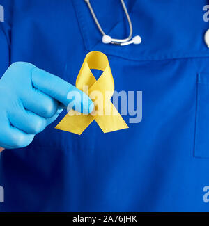doctor in a blue uniform and sterile latex gloves holds a yellow ribbon in his hand, symbol of the fight against the cat's tumor, problem of suicides Stock Photo
