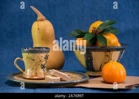 still life of mandarins or oranges with leafs and a pumpkin cookies on a plate next to a cup designed with a floral design against a blue denim backgr Stock Photo