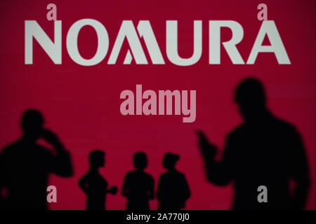 The Nomura logo is seen on an LED screen in the background while a silhouetted person uses a smartphone (Editorial use only) Stock Photo